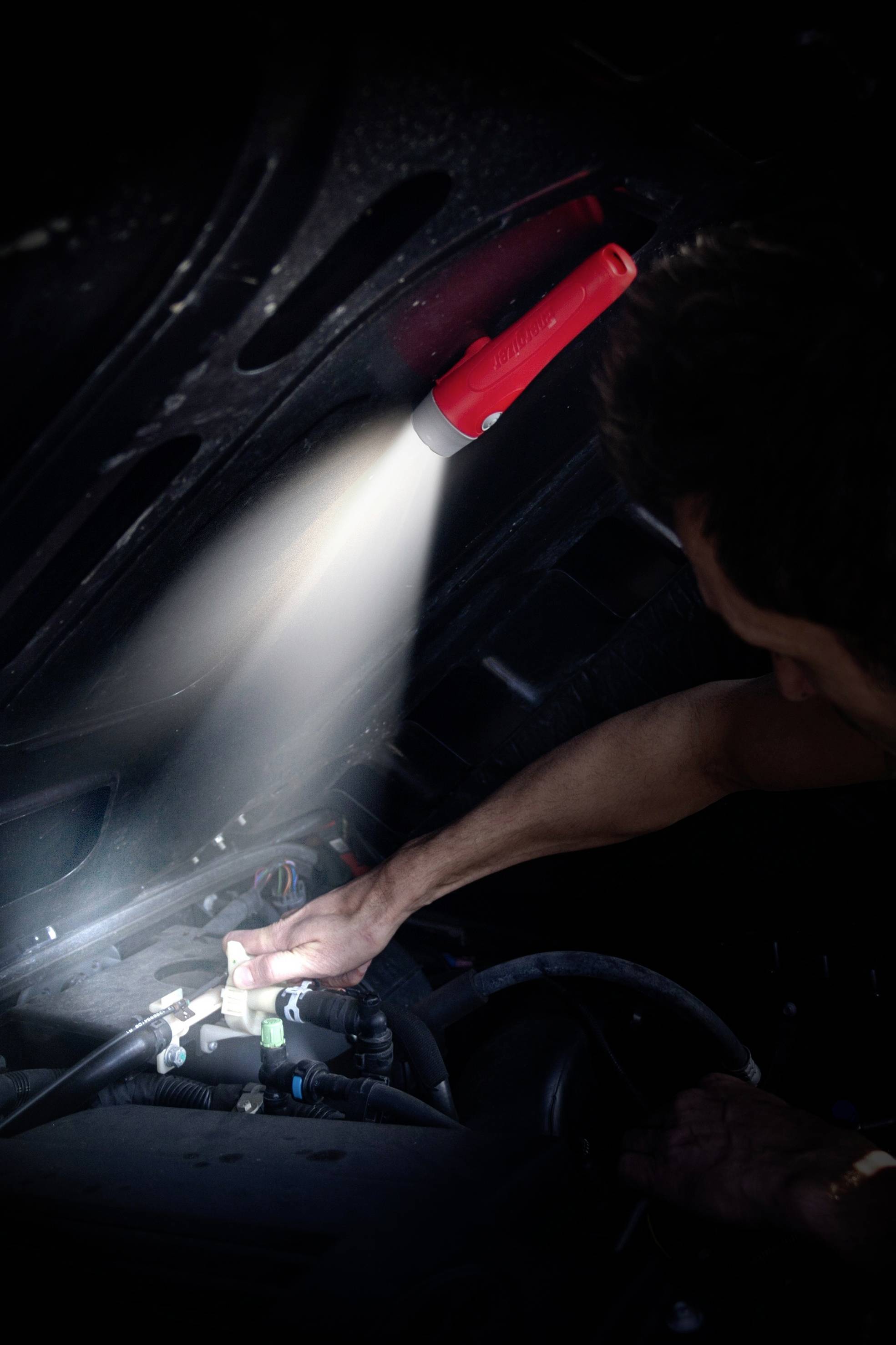 Ein Mechaniker arbeitet mit einer Taschenlampe an einem Automotor. Die Lampe beleuchtet den Motorraum, während der Mechaniker Teile inspiziert.