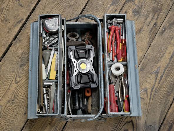 Geöffneter Werkzeugkasten auf Holzboden, voll mit verschiedenen Handwerkzeugen wie Zangen, Schraubenschlüsseln und einem Maßband.