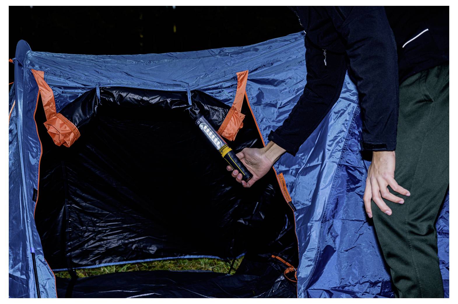 Eine Person mit einer Stirnlampe inspiziert das Innere eines blauen Campingzelts mit offener Tür.
