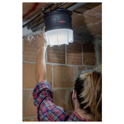 Person in Karihemd greift nach oben, um ein an der Decke montiertes Arbeitslicht in einem unfertigen Ziegelzimmer einzustellen.
