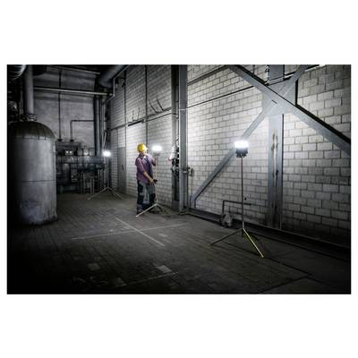 Arbeiter mit Schutzhelm inspiziert Rohre und Ventile in einem düsteren Industriekeller, der von tragbaren Arbeitsleuchten beleuchtet wird.