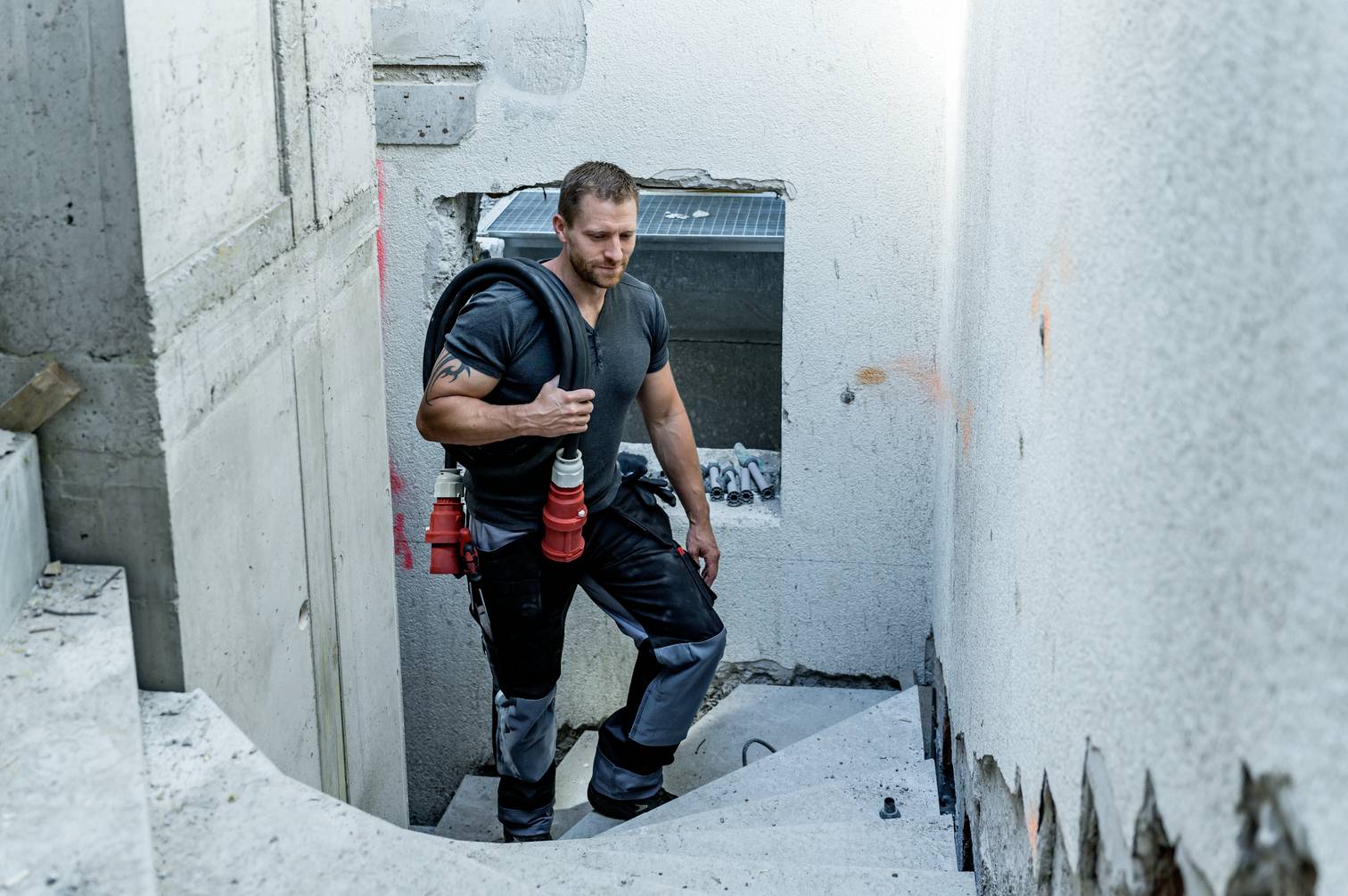 Ein Mann in Arbeitskleidung trägt Equipment über eine Baustellentreppe. Er schaut entschlossen, umgeben von unvollständigen Wänden.