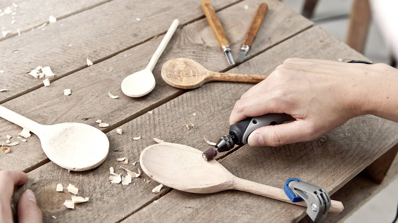 Person schnitzt Holzlöffel mit einem Rotationswerkzeug auf Holztisch. Weitere unfertige Löffel und Holzspäne sind sichtbar.