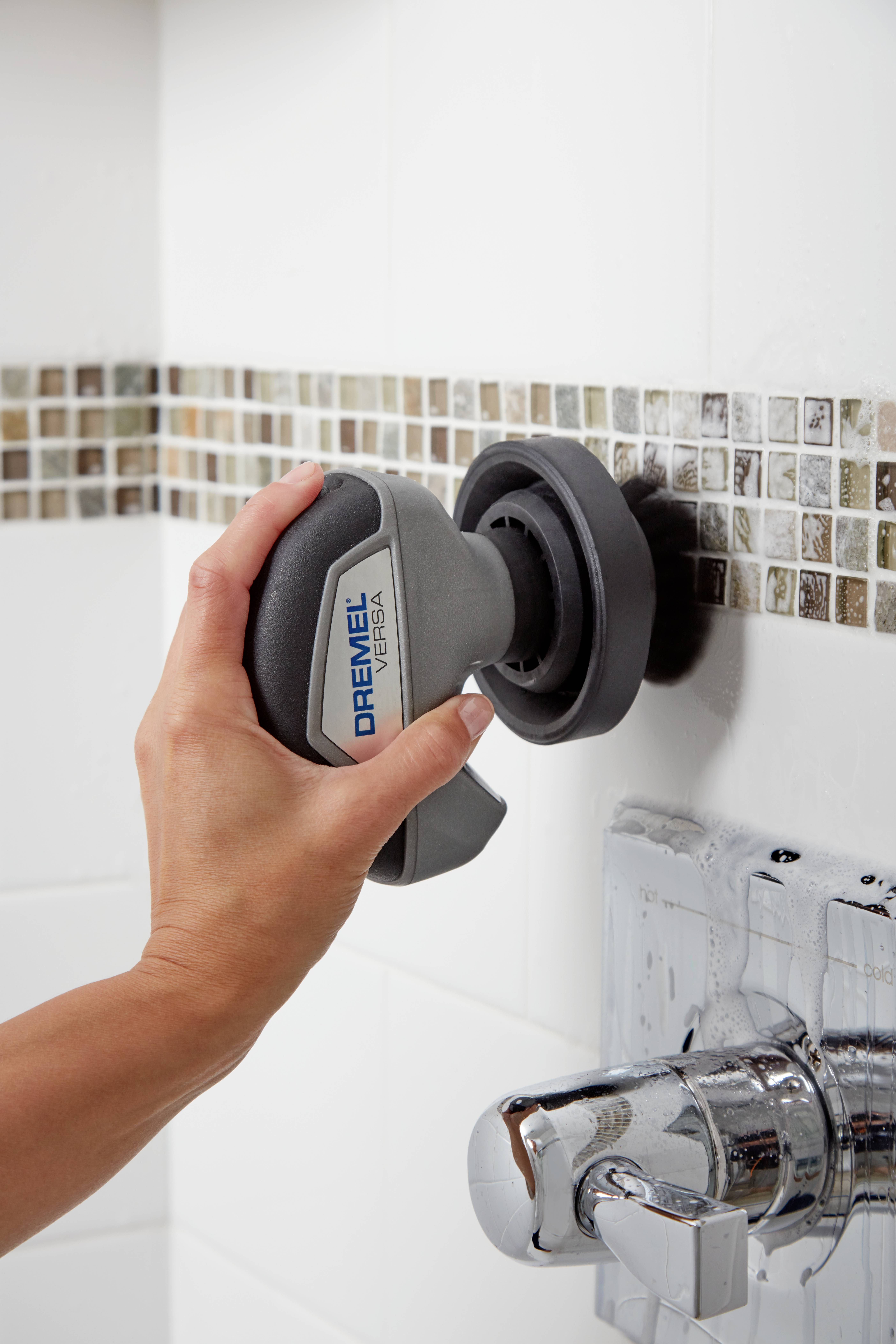 Eine Person reinigt eine geflieste Wand mit einem elektrischen Reinigungsgerät. Ein Wasserhahn ist teilweise sichtbar im Vordergrund.