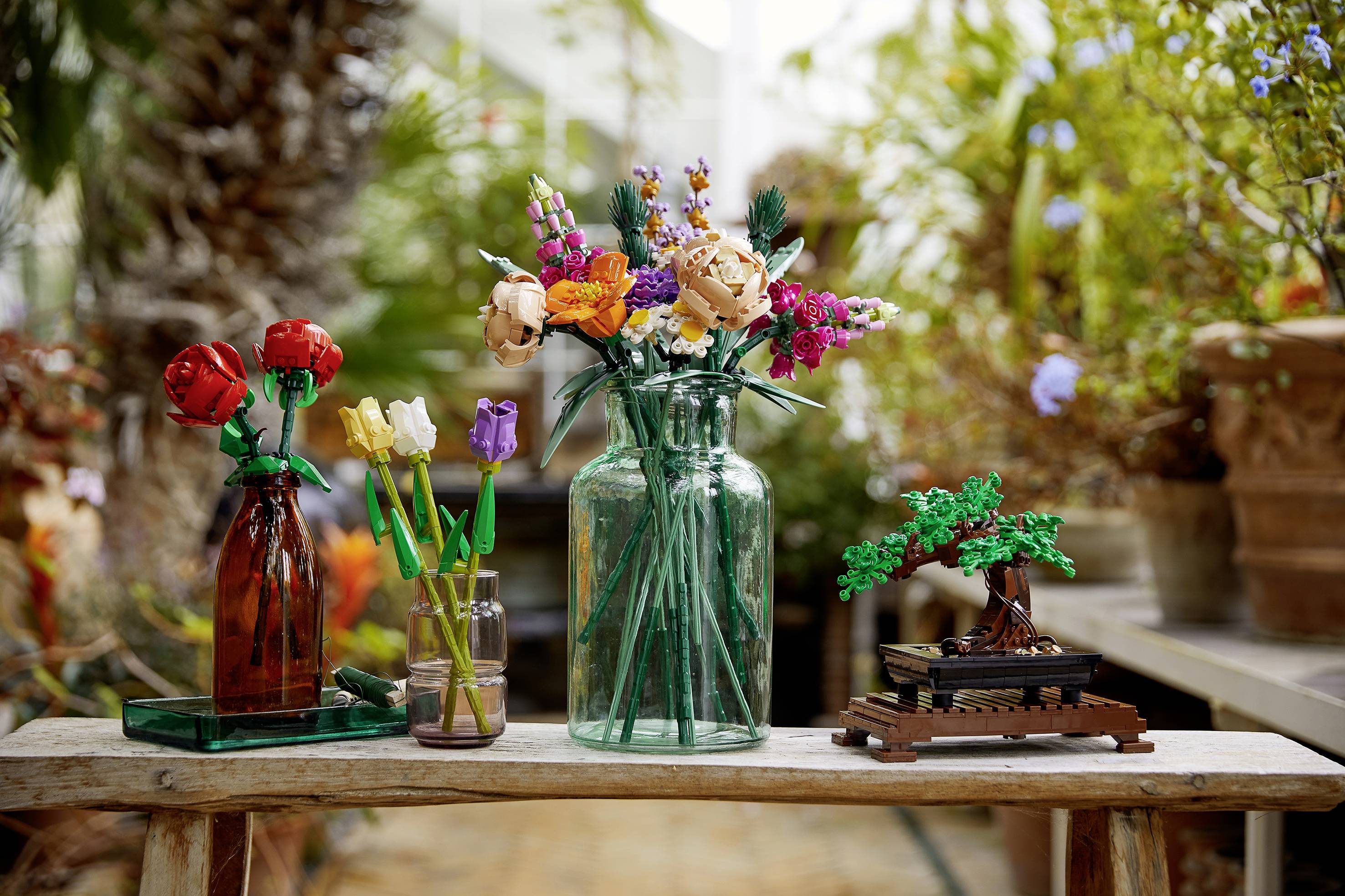 Vasen mit LEGO-Blumen und ein Bonsai-Baum stehen auf einem Tisch in einem botanischen Garten. Im Hintergrund sind Pflanzen zu sehen.