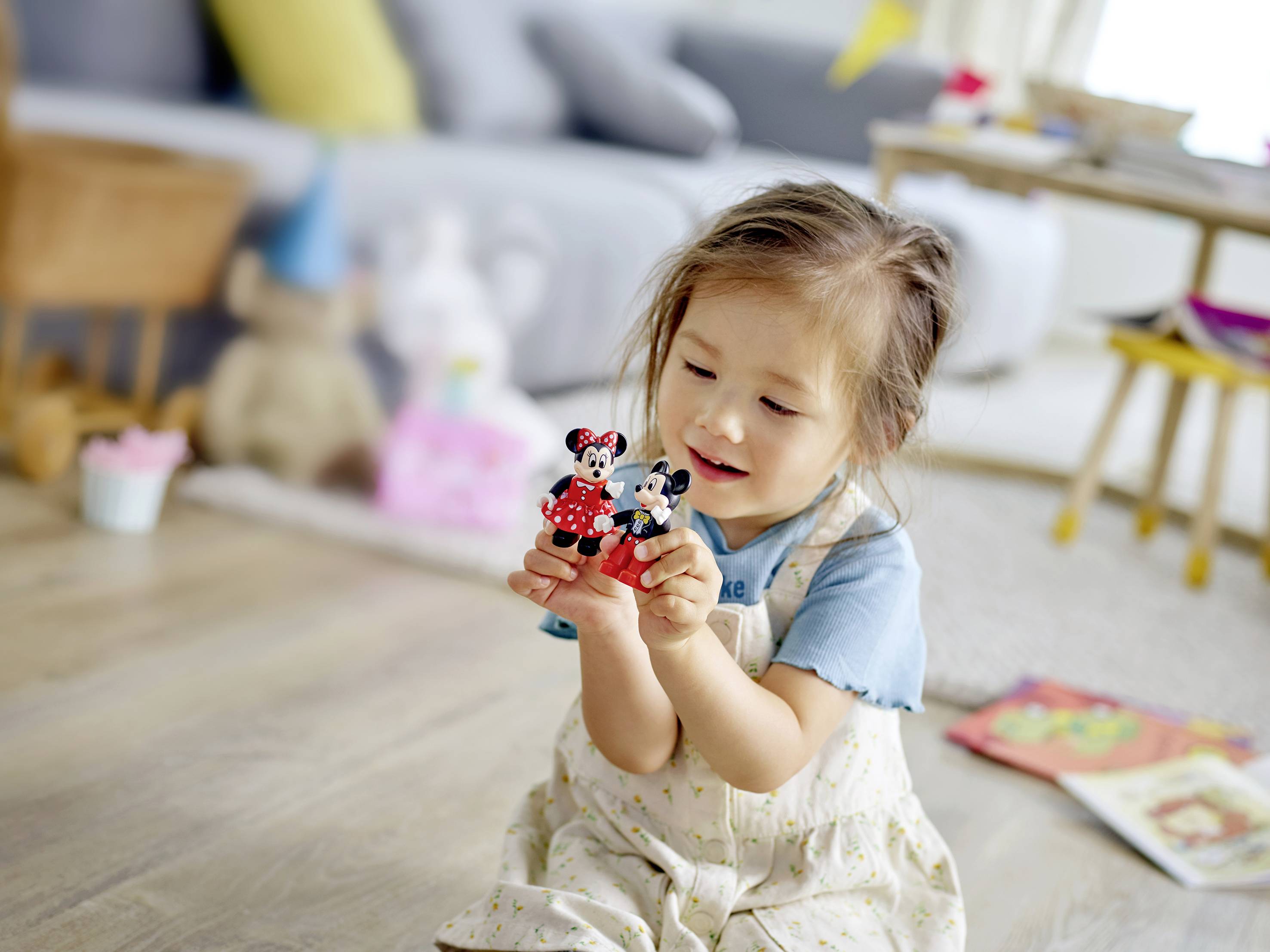 Ein kleines Mädchen sitzt auf dem Boden und hält zwei Spielzeugfiguren, eine in jedem Hand. Im Hintergrund unscharfe Wohnzimmermöbel.