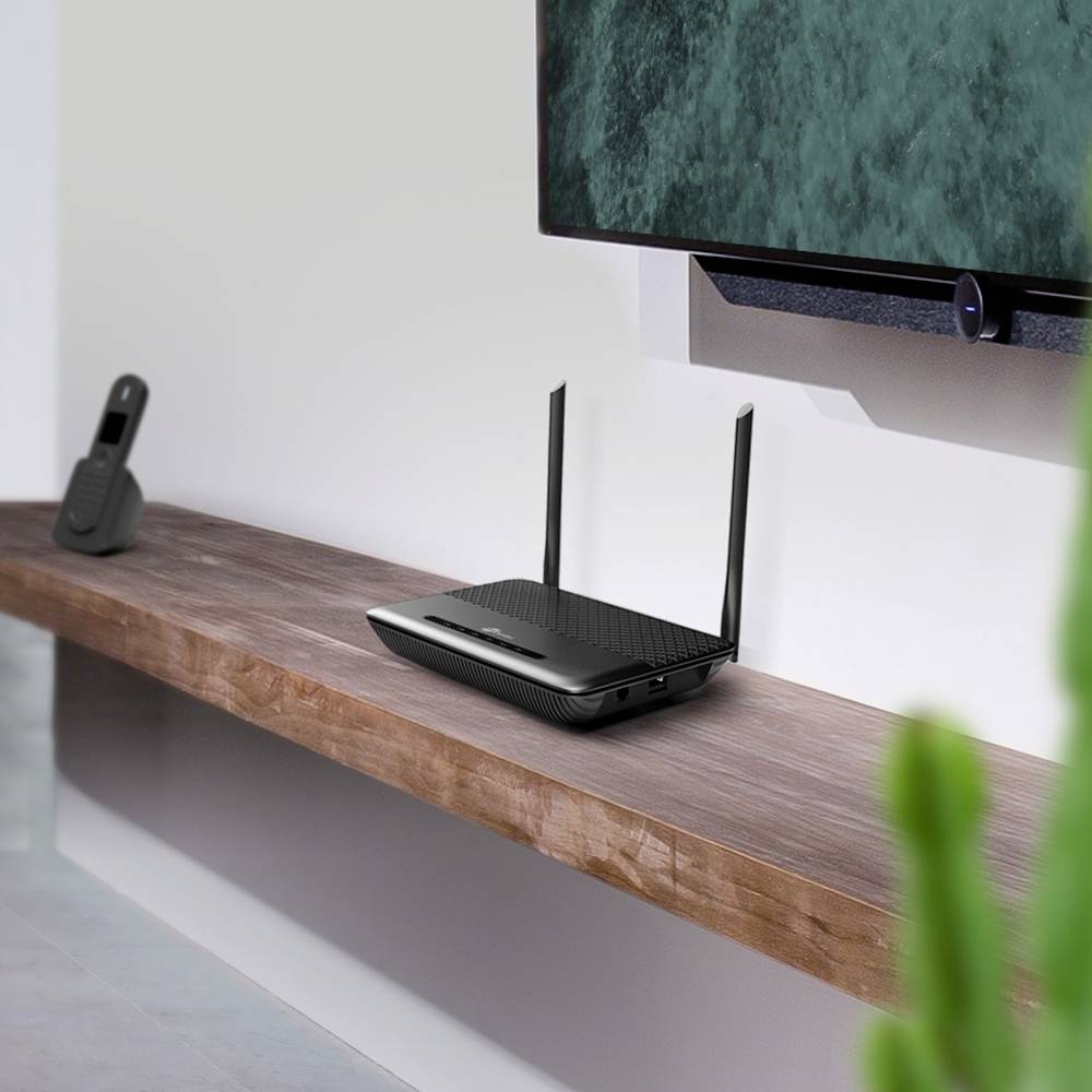Internet-Router mit Antennen auf einem Holztisch vor einer weißen Wand. Im Hintergrund ein Fernsehbildschirm, an der Wand montiert.