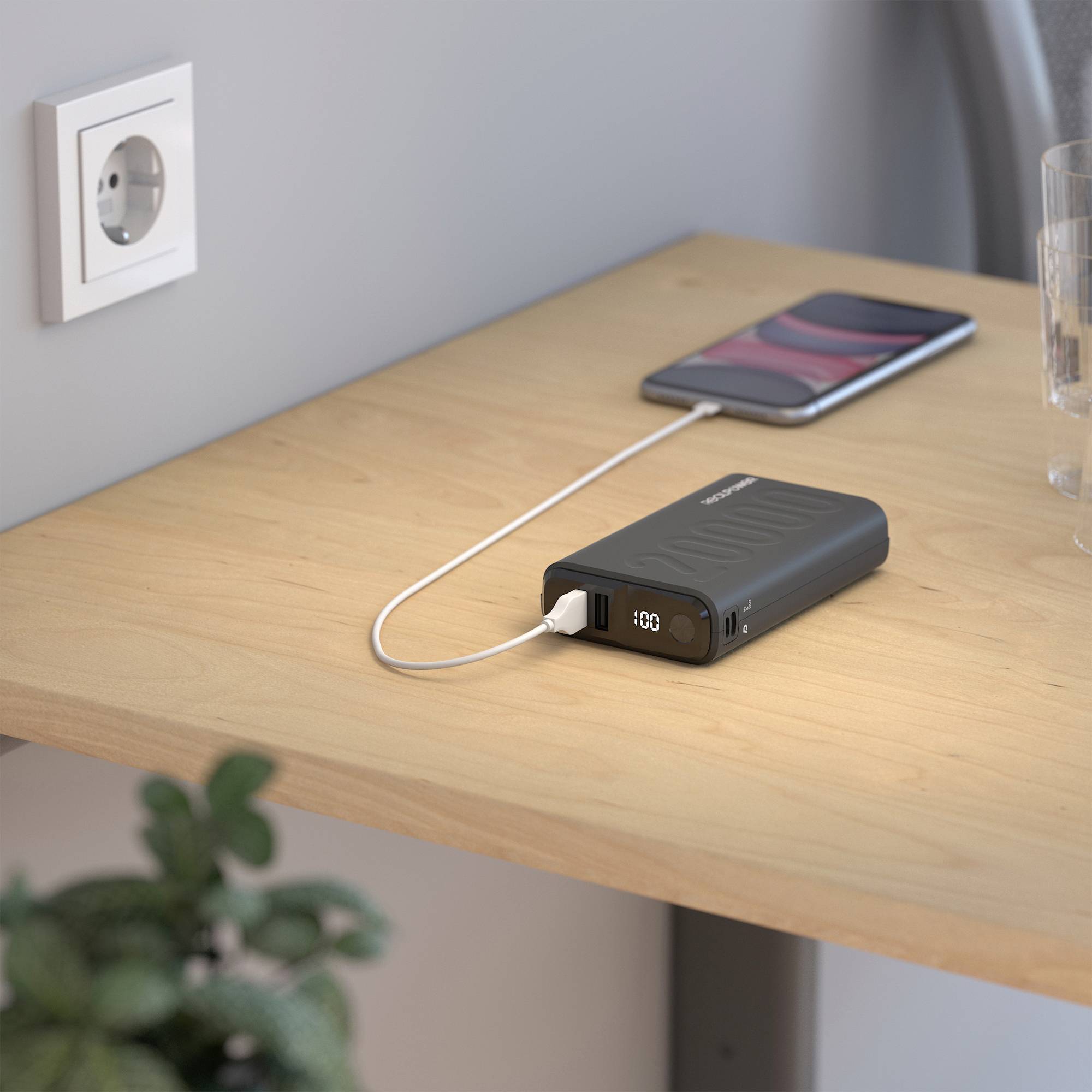 Ein Smartphone wird auf einem Holztisch mit einem tragbaren Ladegerät über ein Ladekabel geladen; eine Steckdose an der Wand.