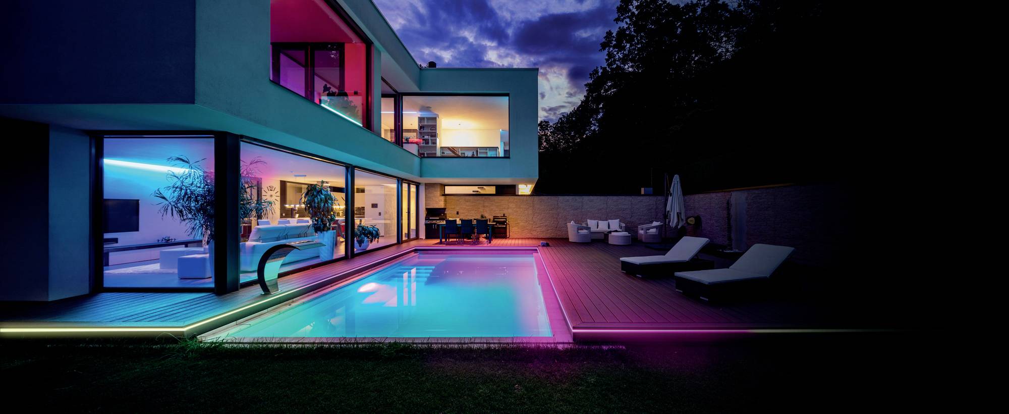 Ein modernes Haus bei Nacht mit beleuchtetem Pool und neonfarbenen Lichtern in den Fenstern, umgeben von Liegestühlen und einem Tisch.
