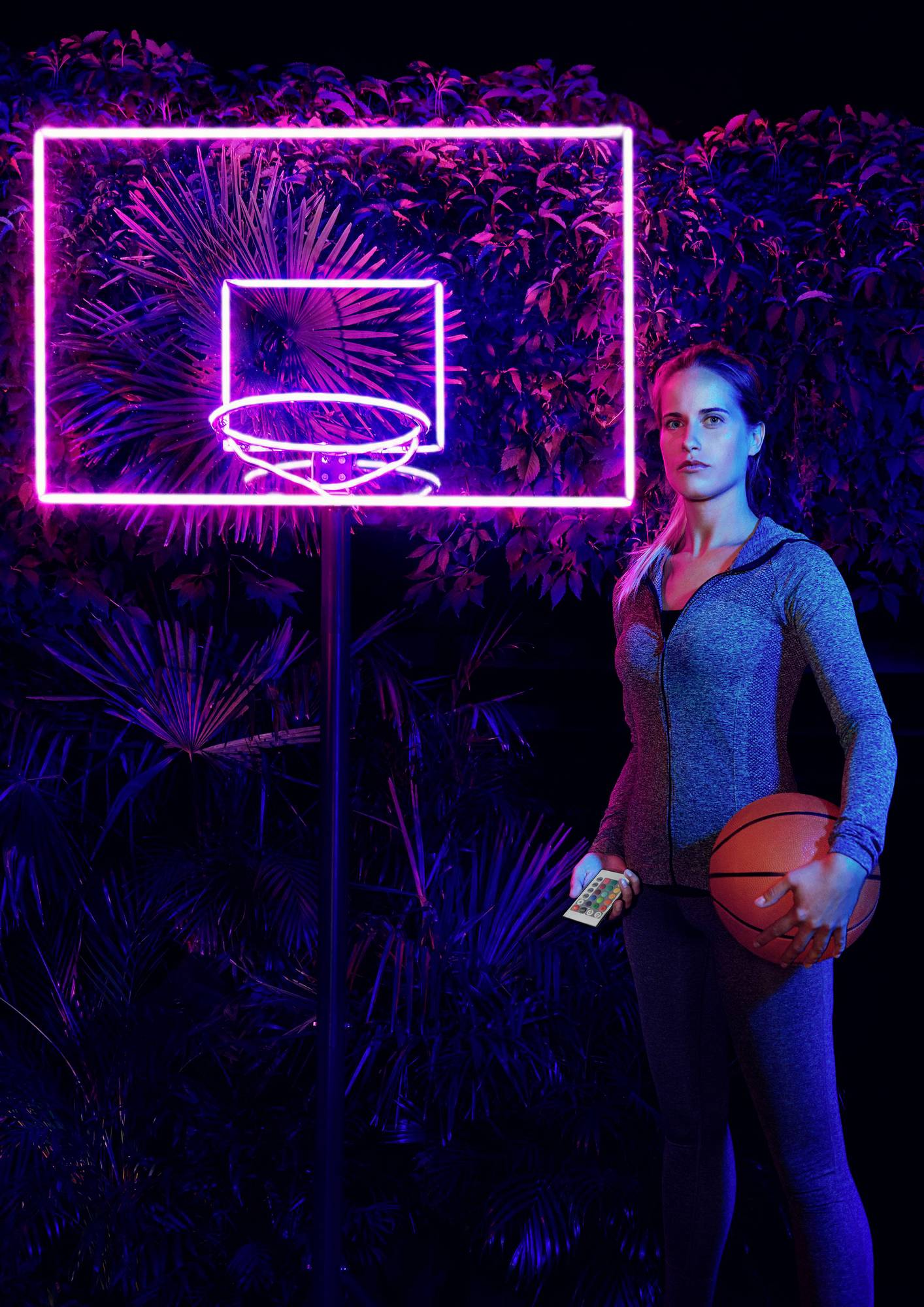 Eine Frau im Sportanzug hält einen Basketball und steht vor einem neonbeleuchteten Basketballkorb. Im Hintergrund sind Pflanzen sichtbar.
