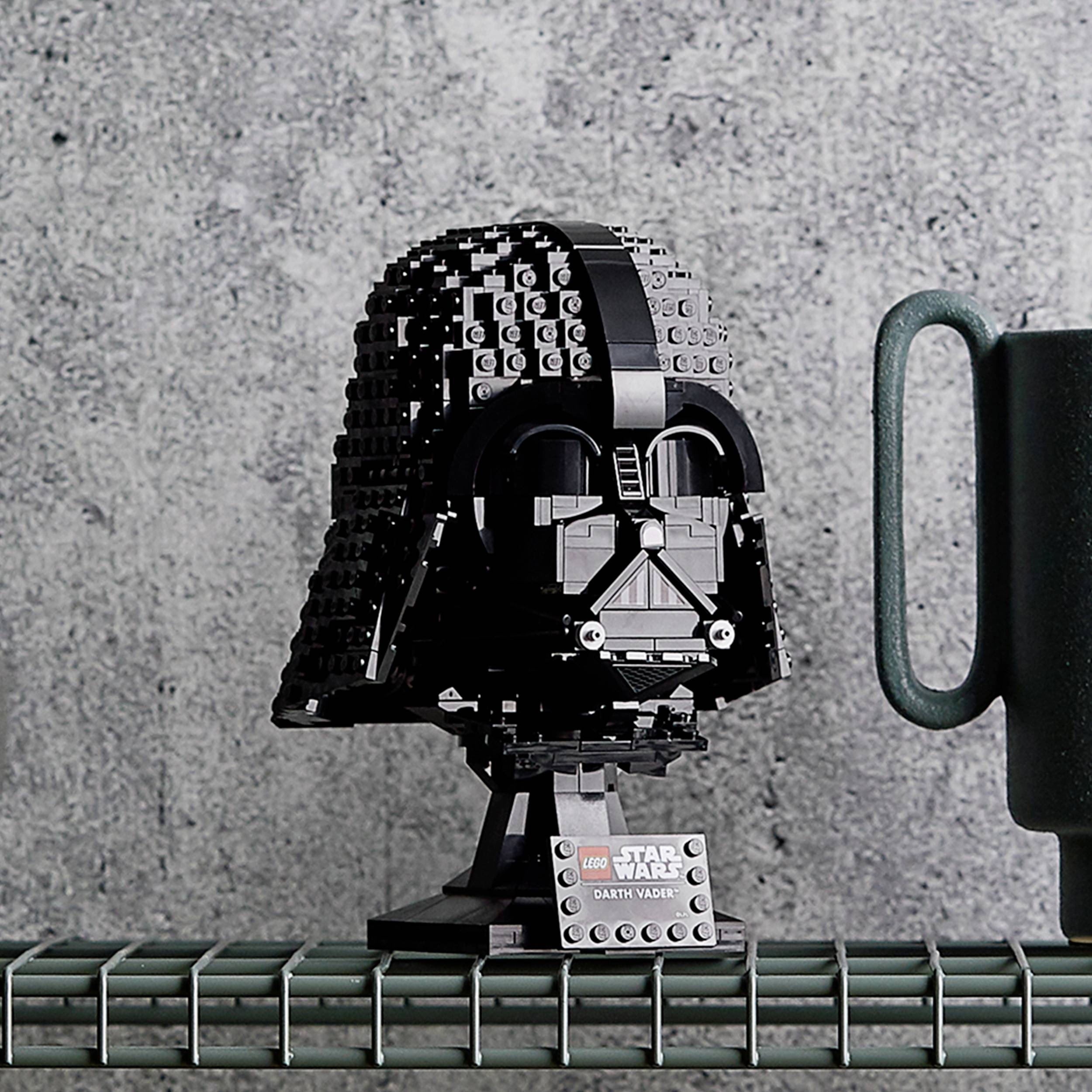 Ein LEGO-Modell eines bekannten schwarzen Helms und ein grüner Becher stehen auf einem Gitterregal vor einer grauen Wand.