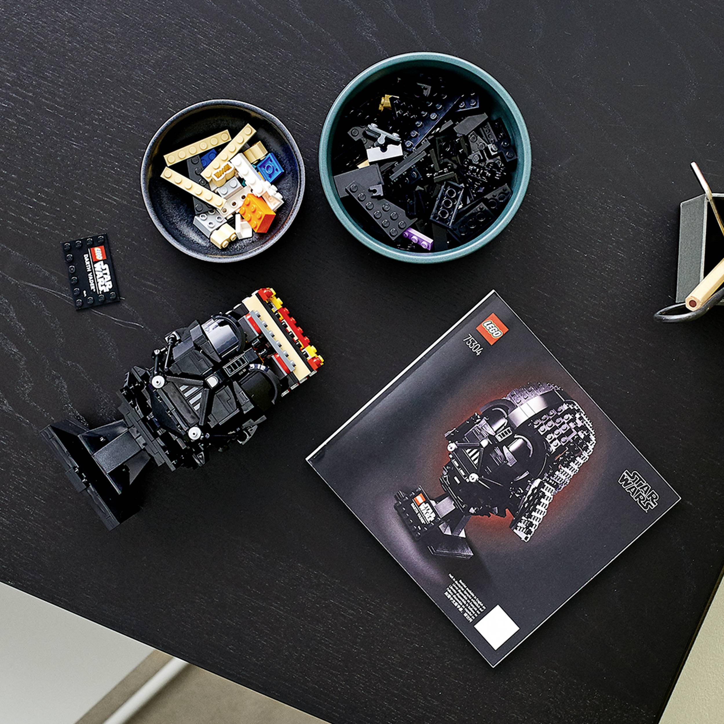 Lego-Set zum Bau eines schwarzen Helms liegt auf einem Tisch, umgeben von zwei Schüsseln mit Legosteinen und einer Bauanleitung.