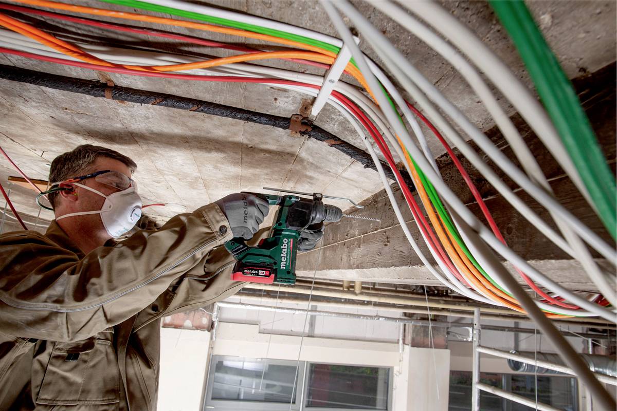 Ein Handwerker bohrt in eine Decke und trägt dabei Schutzkleidung und eine Maske. Über ihm sind bunte Kabel verlegt.