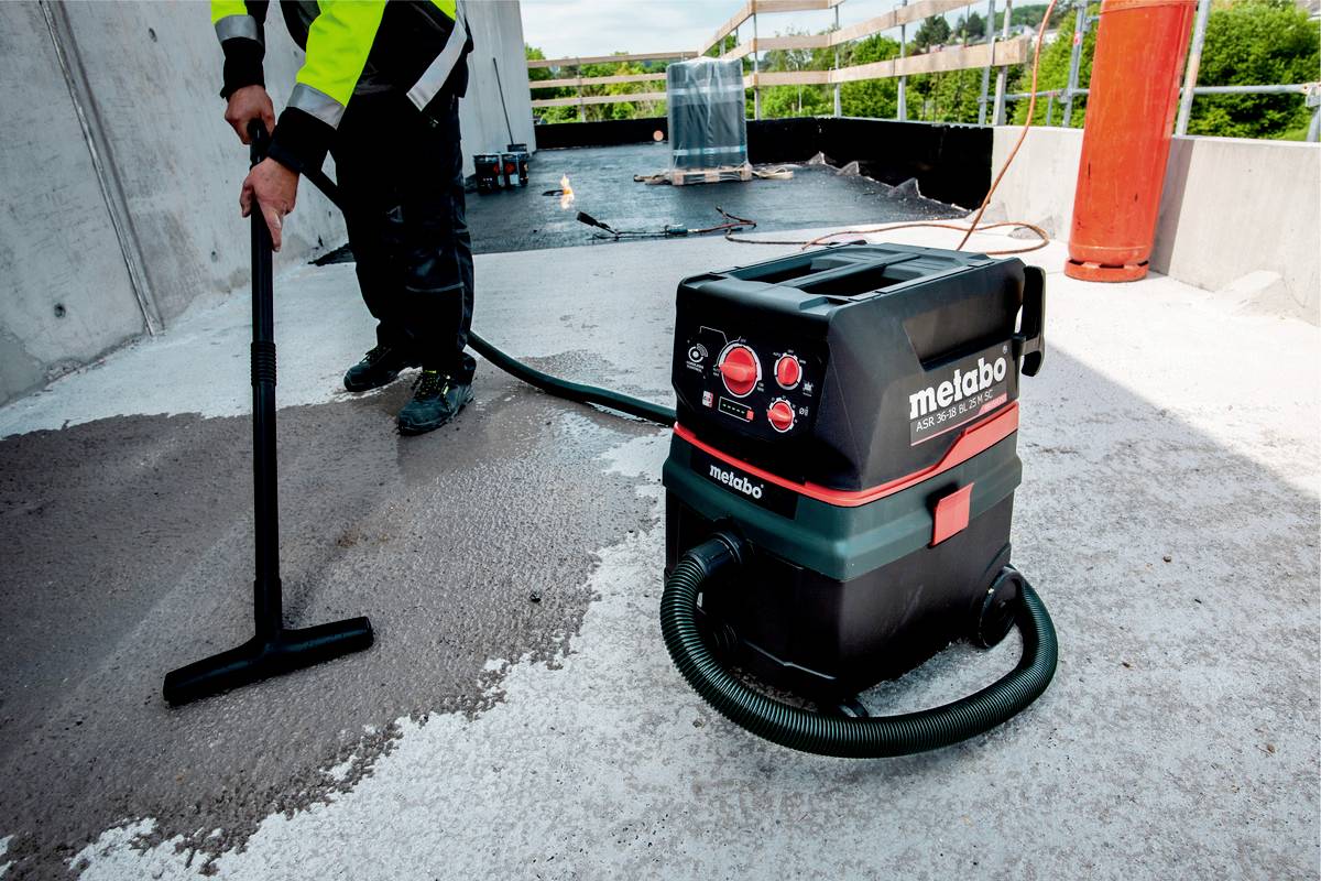 Ein Arbeiter reinigt eine Baustelle mit einem Industriestaubsauger. Der Staubsauger steht im Vordergrund, während der Arbeiter im Hintergrund arbeitet.