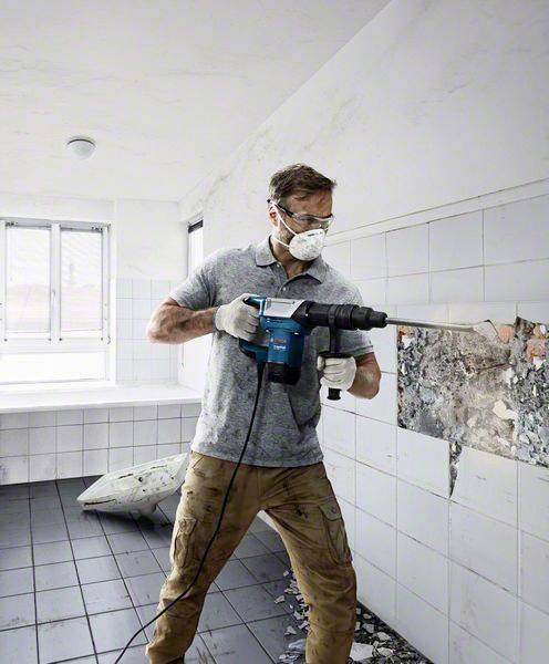 Ein Mann mit Schutzbrille und Maske nutzt einen Elektrowerkzeug-Bohrhammer, um Fliesen von einer Wand in einem Raum zu entfernen.