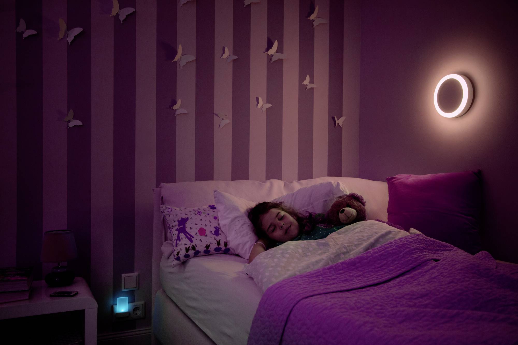Kind schläft friedlich im Bett, umgeben von Kuscheltieren, in einem lila beleuchteten Zimmer mit Wanddekorationen von Vögeln.
