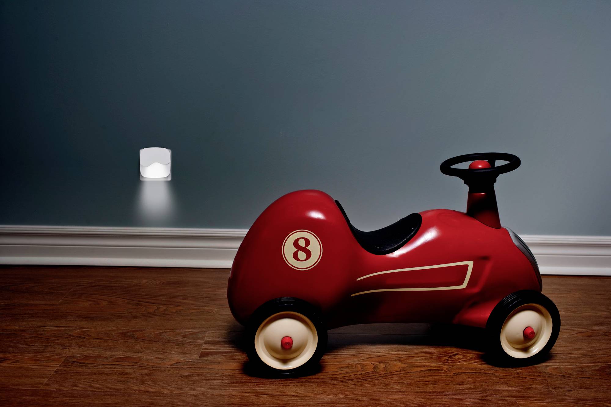 Ein rotes Spielzeugauto mit der Nummer 8 steht auf einem Holzboden vor einer blauen Wand. Im Hintergrund leuchtet ein Nachtlicht.