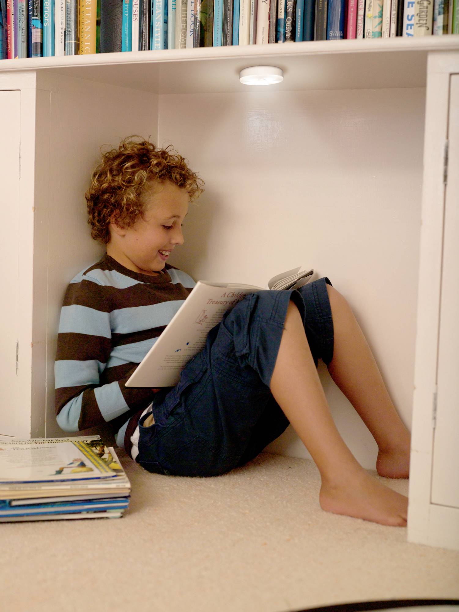 Ein Junge sitzt lächelnd in einer Ecke unter einem Bücherregal und liest ein Buch. Stapel von Büchern liegen neben ihm.