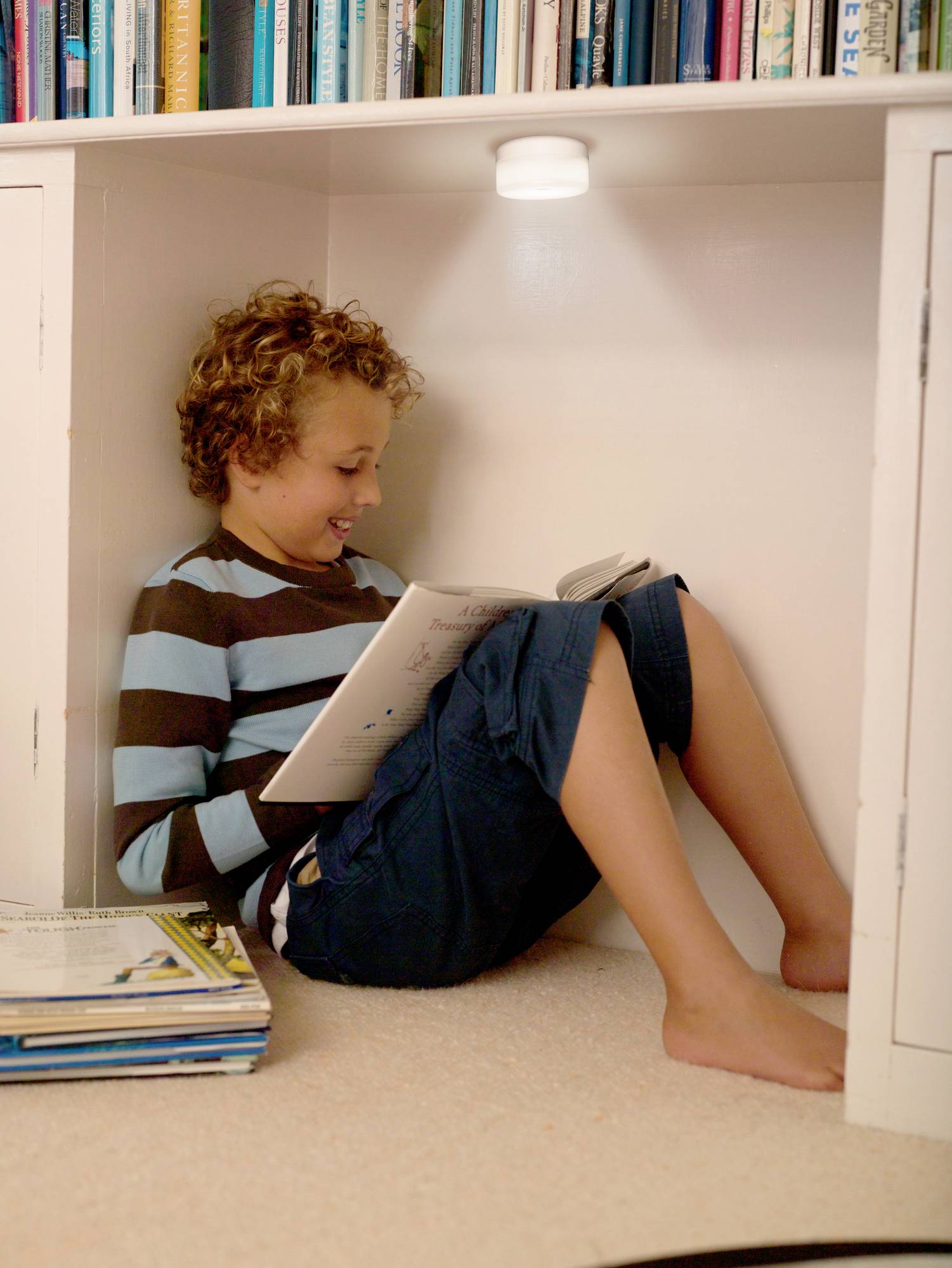 Ein Kind sitzt lächelnd unter einem Bücherregal und liest gemütlich ein Buch. Im Hintergrund sind mehrere Bücher zu sehen.