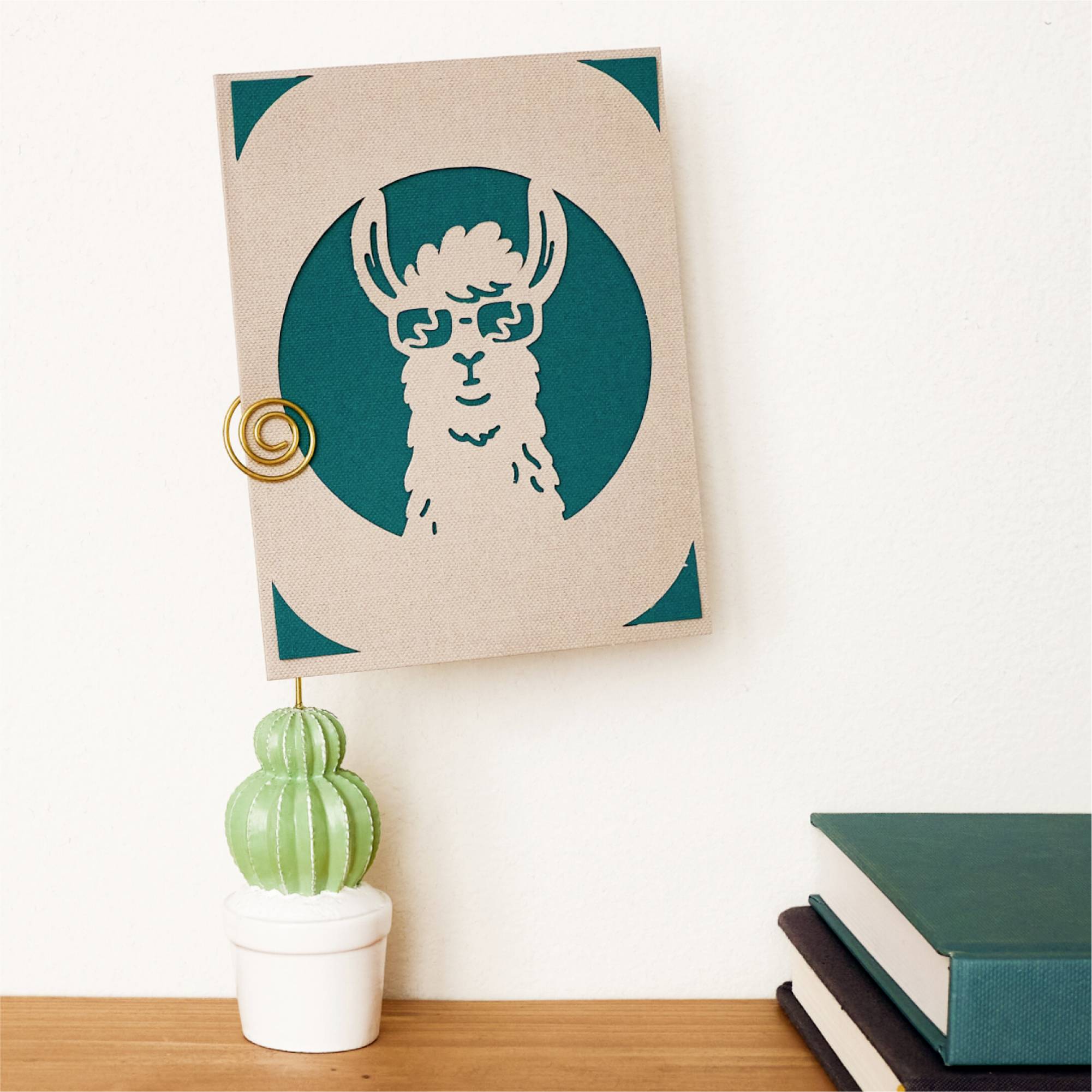 Eine Karte mit dem Bild eines stilisierten Lamas mit Brille steht aufrecht in einem Mini-Kaktushalter. Daneben liegen Bücher.
