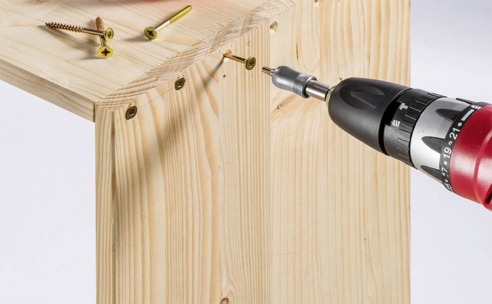 Eine elektrische Bohrmaschine zieht Schrauben in ein Holzstück ein, während einige Schrauben darauf liegen.