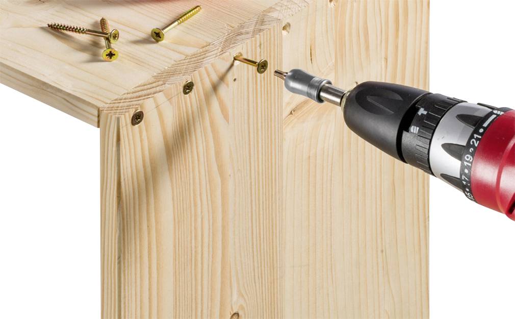 Ein Akkubohrer zieht eine Schraube in einem Holzwinkel fest. Mehrere Schrauben sind auf der Oberfläche verteilt.