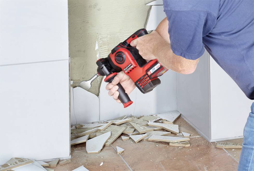 Eine Person verwendet eine rote Bohrmaschine, um Fliesen von einer Wand zu entfernen. Fliesenreste liegen auf dem Boden.