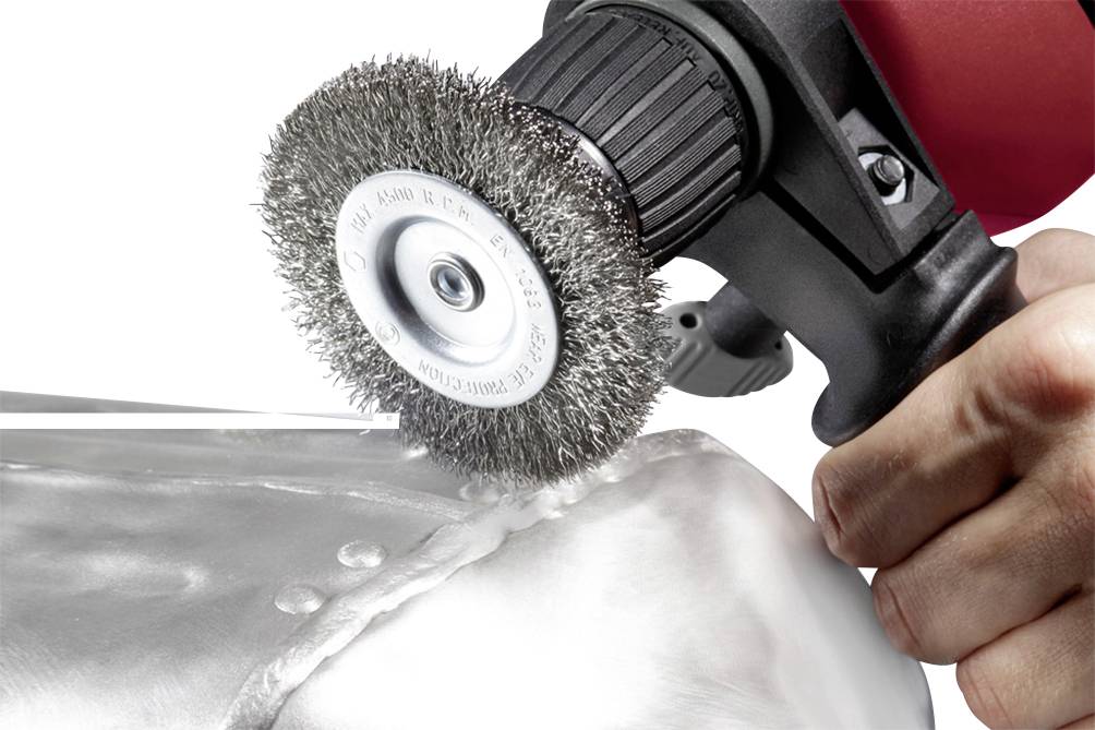 Eine Hand benutzt eine elektrische Drahtbürste, um eine Metalloberfläche, möglicherweise eine Schweißnaht, zu schleifen oder zu reinigen.