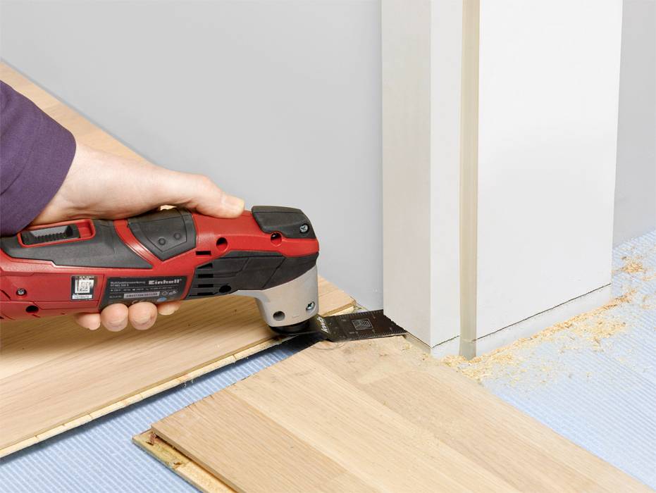 Eine Hand hält ein elektrisches Multifunktionswerkzeug und schneidet Holz an einer Türzarge. Sägespäne sind sichtbar auf dem Boden.