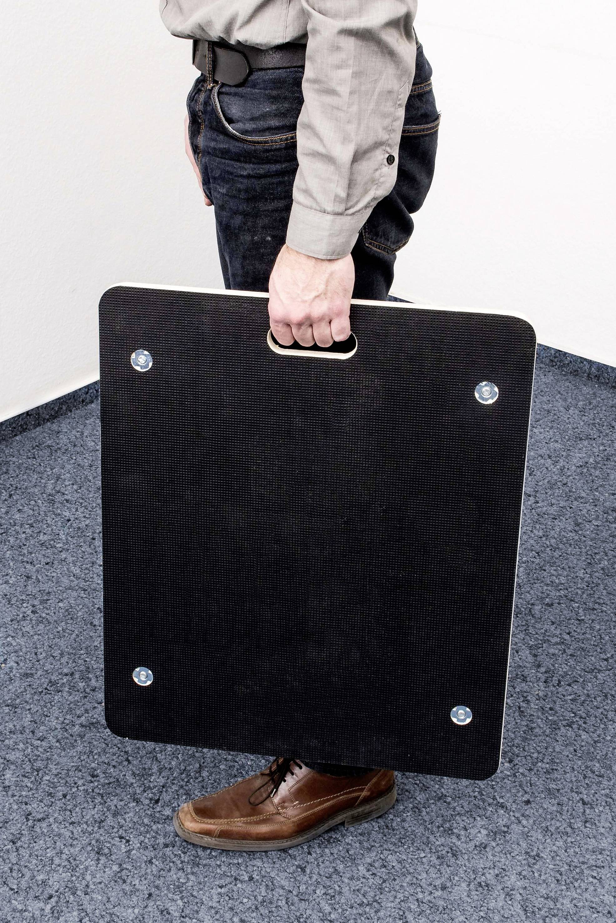 Eine Person hält eine große, rechteckige, schwarze Tafel mit einer integrierten Grifföffnung in der rechten Hand, stehend auf blauem Teppichboden.