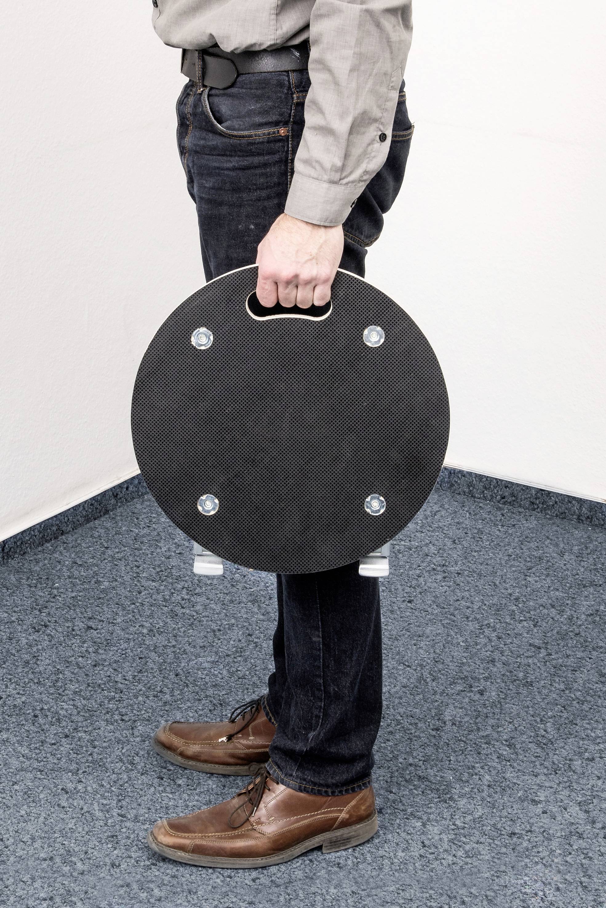 Person steht auf Teppichboden und hält eine runde, schwarze Platte mit Griff in der Hand. Die Platte hat vier silberne Schrauben.
