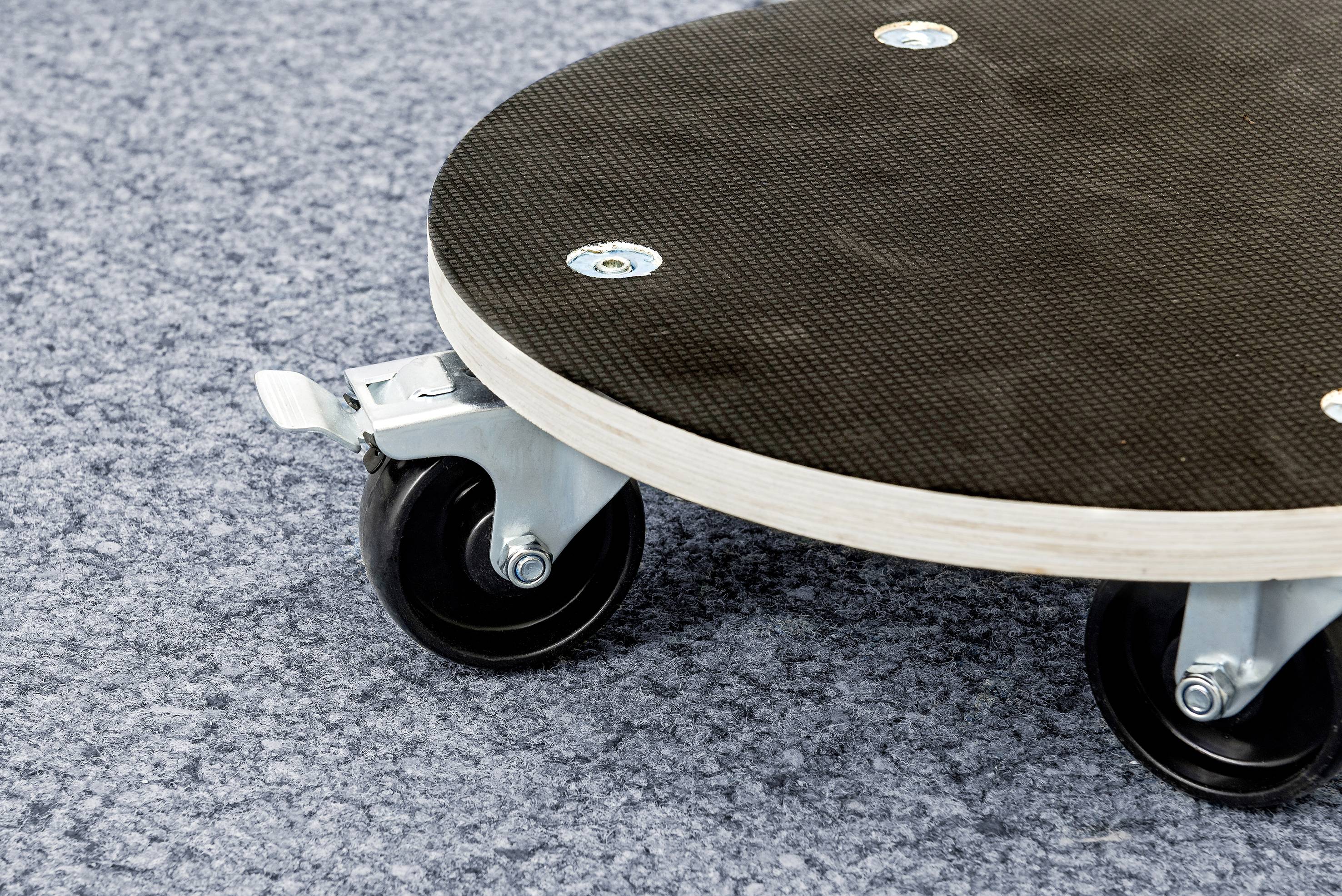 Eine runde schwarze Plattform mit Rollen auf grauem Teppichboden. Sie wird möglicherweise als Transporthilfe genutzt.