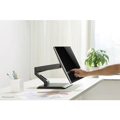 Ein Finger berührt den Bildschirm eines modernen, verstellbaren Monitors auf einem Schreibtisch neben Schreibmaterialien und einer Pflanze.