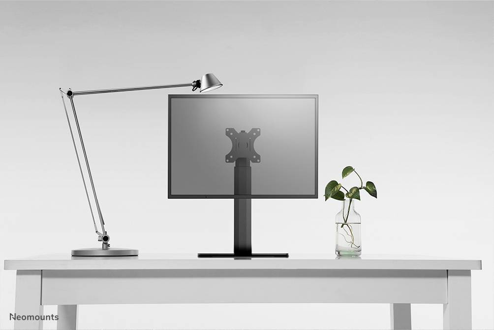 Ein Schreibtisch mit einer modernen Lampe links, einem Monitor in der Mitte und einer Pflanze in einem Glasgefäß rechts.