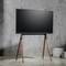 Ein moderner Fernseher auf einem dreibeinigen Holzständer steht in einem minimalistischen Raum mit Betonwänden.