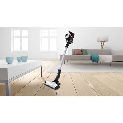 Kabelloser Staubsauger steht auf Holzfußboden in modernem Wohnzimmer mit Sofa, Tisch und Lampe. Helle und minimalistische Einrichtung.