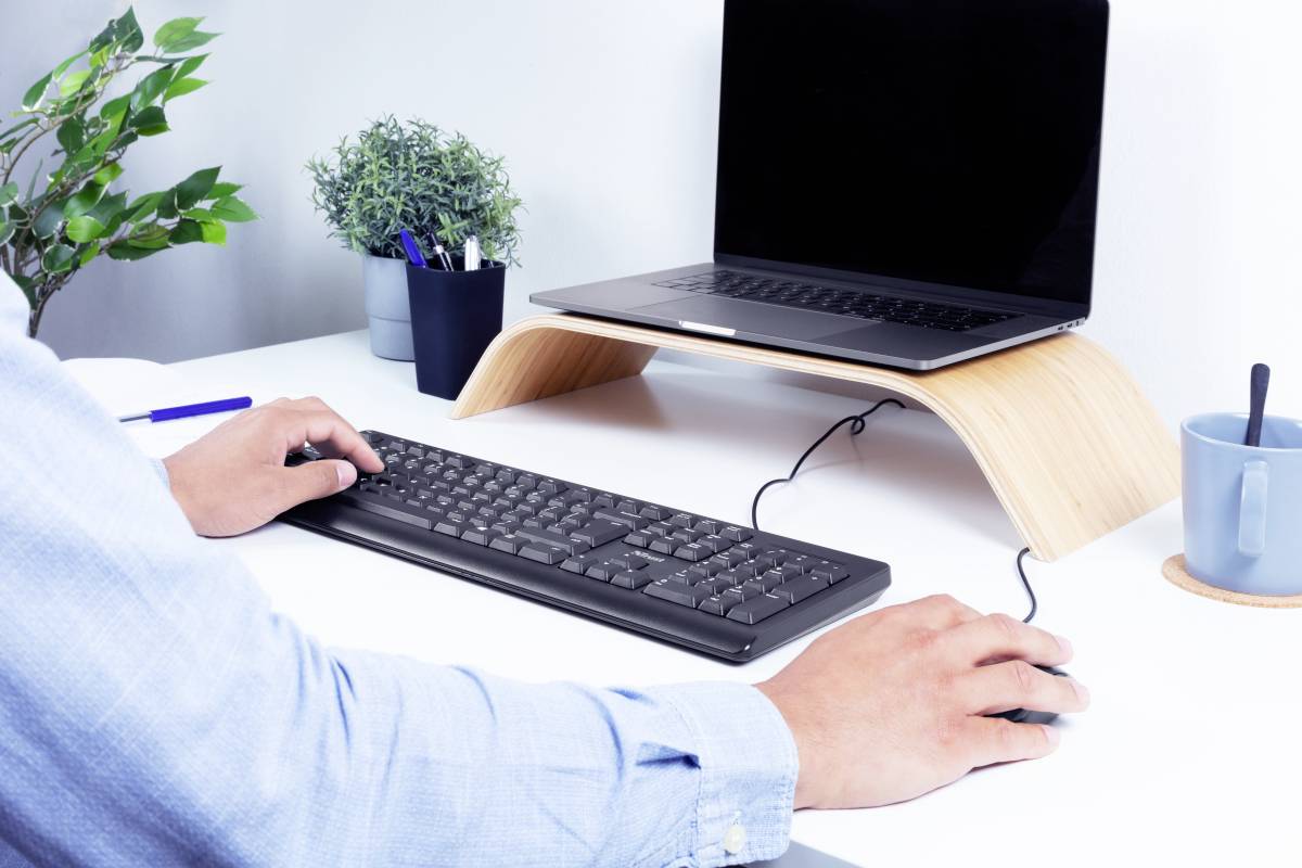 Person arbeitet an einem Schreibtisch mit Laptop, Tastatur und Maus. Der Laptop steht auf einem Holzständer. Eine Pflanze und Tasse stehen daneben.