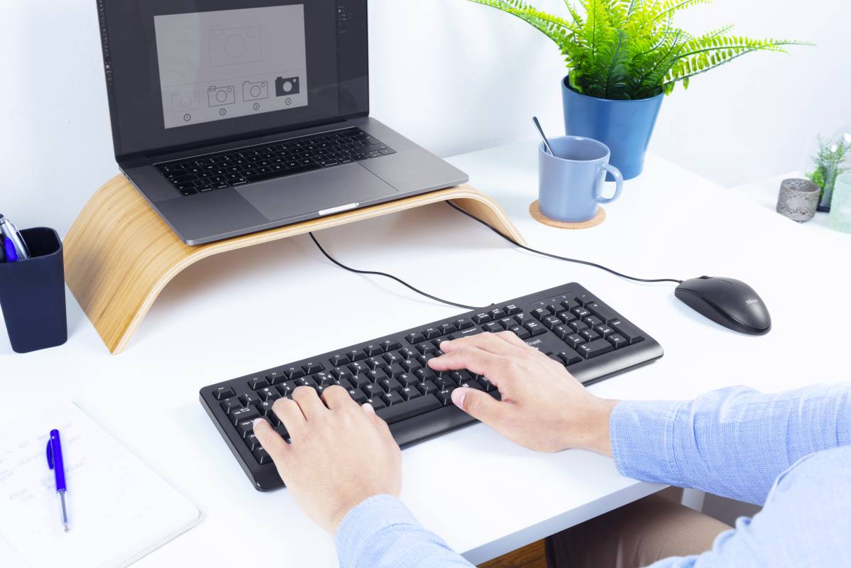 Hände tippen auf einer Computertastatur auf einem weißen Schreibtisch. Im Hintergrund ein Laptop, eine Tasse und eine Pflanze.