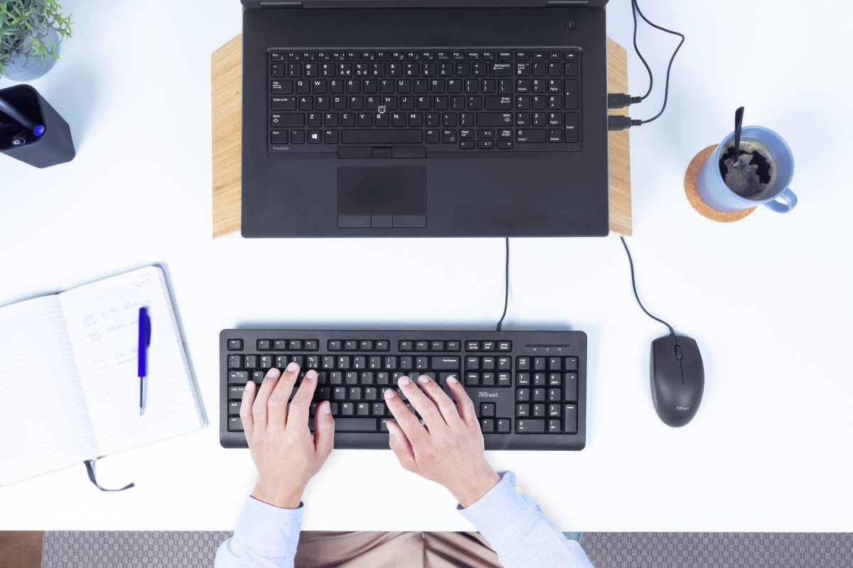 Eine Person tippt an einem externen Keyboard, das mit einem Laptop verbunden ist. Daneben liegen eine Maus, ein Notizbuch und eine Tasse Kaffee.