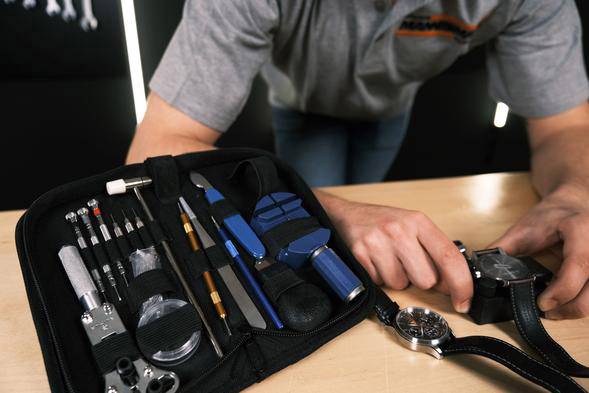 Eine Person arbeitet an einer Uhr an einem Tisch. Daneben liegt ein offenes Reparatur-Set mit verschiedenen Uhrenwerkzeugen.