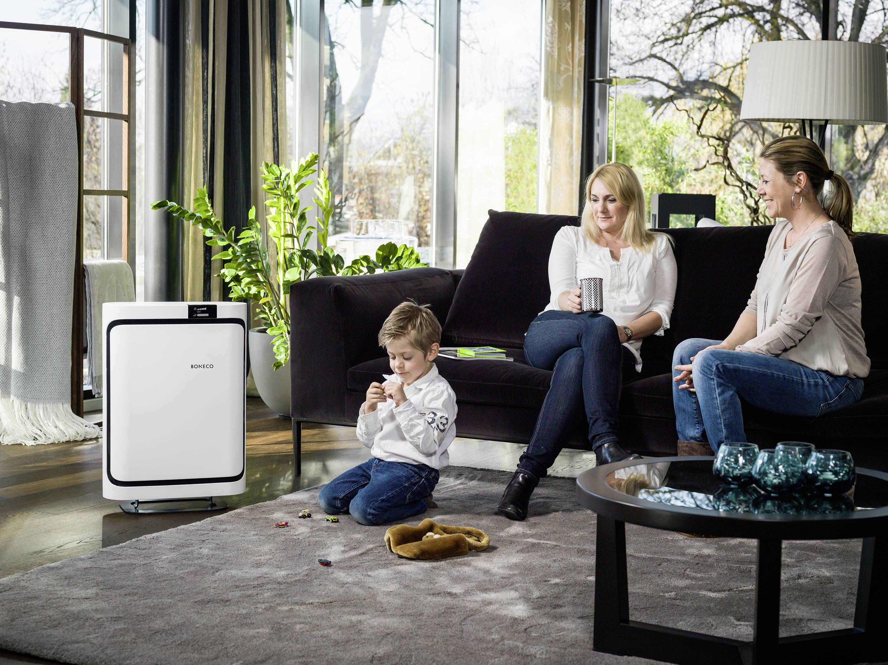 'Ein Kind spielt auf dem Boden, während zwei Erwachsene auf einem Sofa sitzen und Kaffee trinken. Neben ihnen steht ein Luftreiniger.'