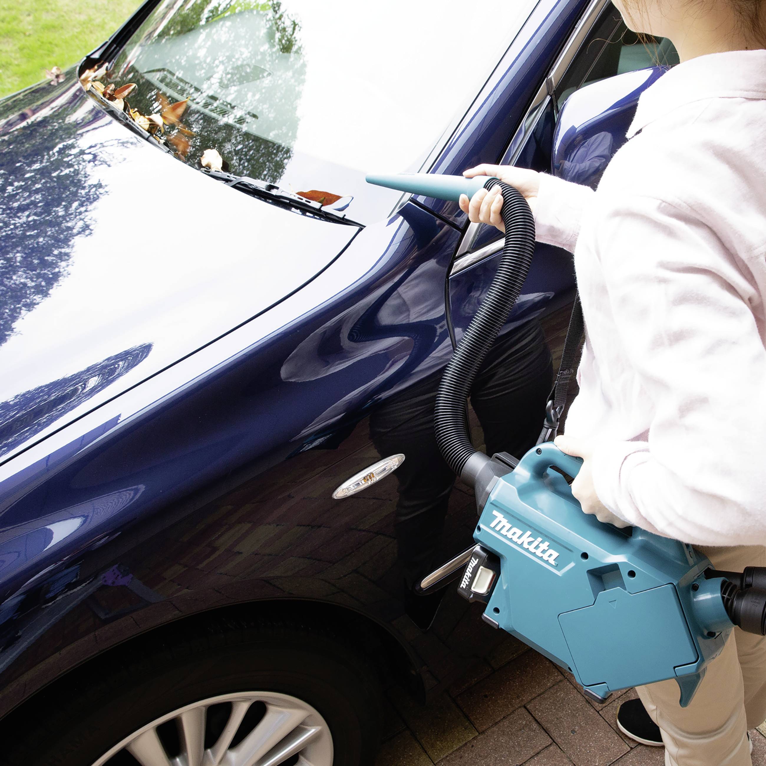Eine Person benutzt einen blauen Staubsauger zum Reinigen eines Autodachs. Das Auto ist schwarz und steht auf einer gepflasterten Fläche.