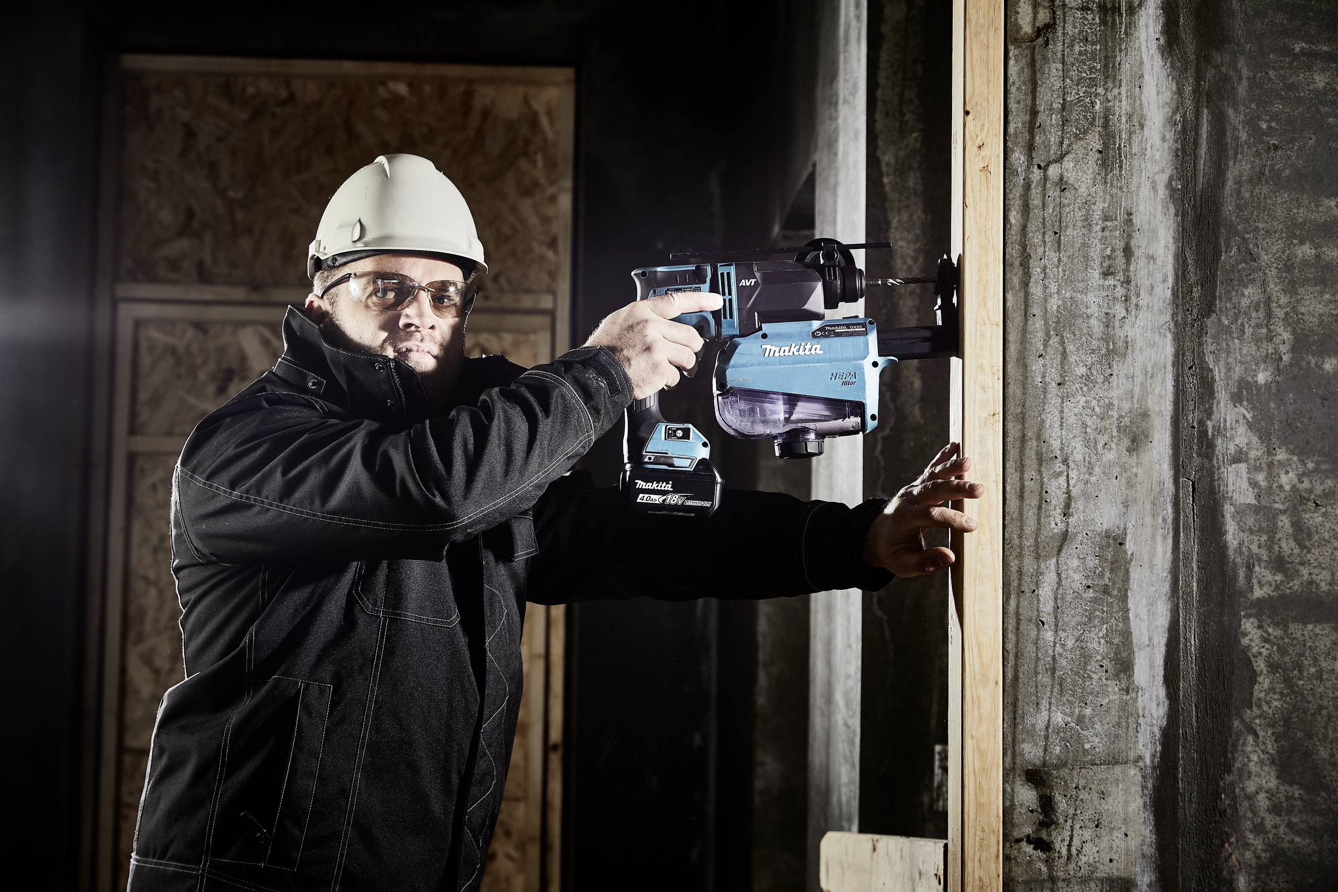 Mann mit Schutzhelm und Brille bohrt mit einer blauen Bohrmaschine in eine Betonwand. Baustellenszene mit Holzrahmen im Hintergrund.