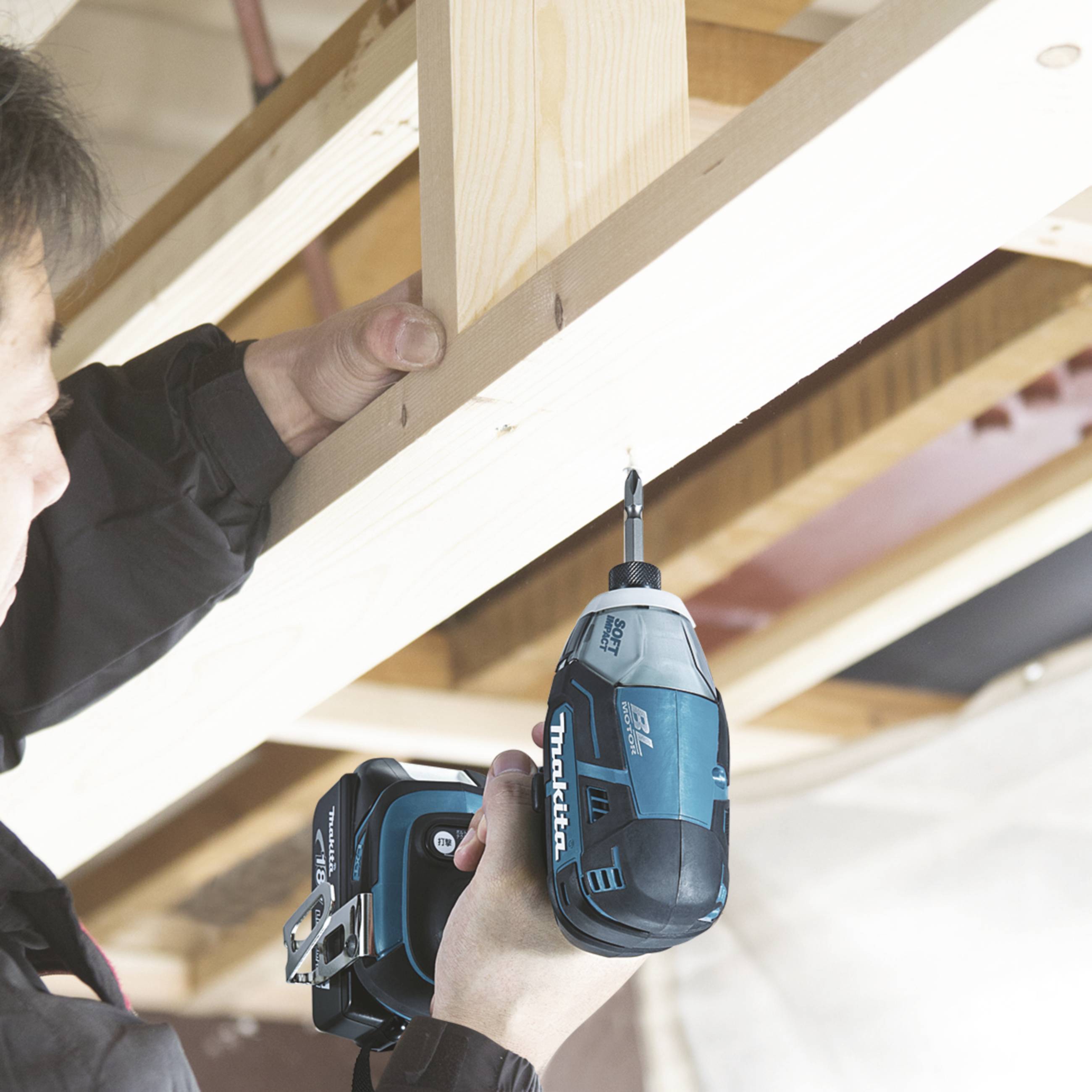 Eine Person verwendet eine blaue Bohrmaschine, um ein Holzbrett an einer Decke zu befestigen. Im Hintergrund sind weitere Holzbalken zu sehen.