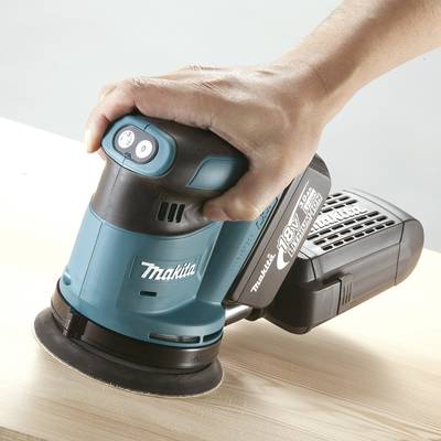 Eine Hand hält einen blauen Schleifer der Marke Makita, während er über eine Holzoberfläche betrieben wird.
