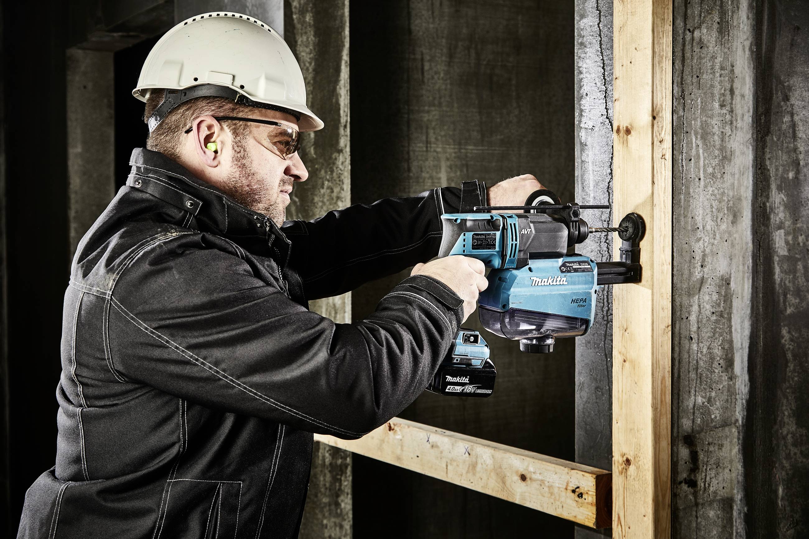 Ein Arbeiter trägt einen Schutzhelm und Ohrstöpsel, während er mit einem Akku-Bohrhammer ein Holzbrett an einer Betonwand befestigt.