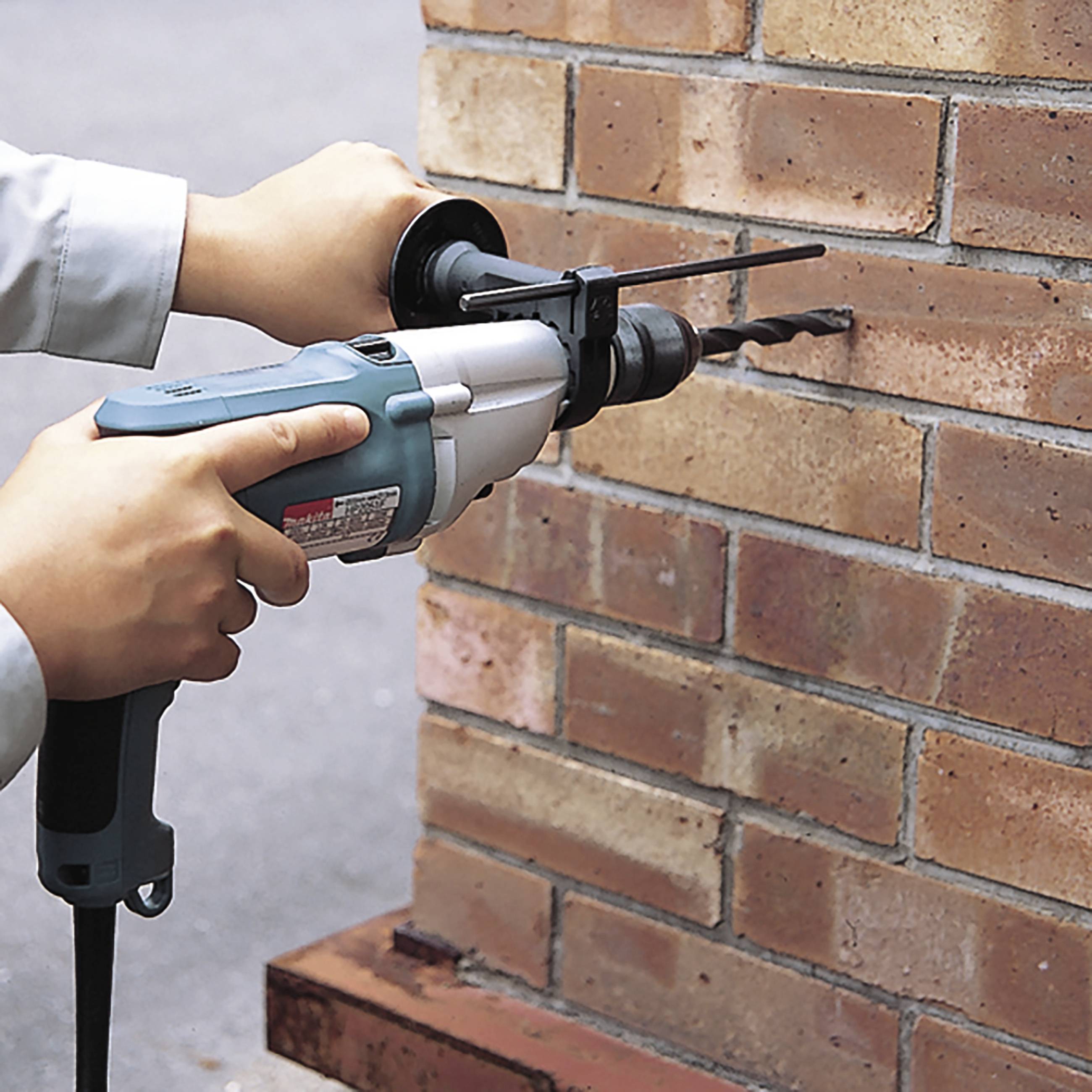 Eine Person benutzt einen Bohrhammer, um in eine Ziegelwand zu bohren. Der Bohrer ist blau und silbern. Es wird im Freien gearbeitet.