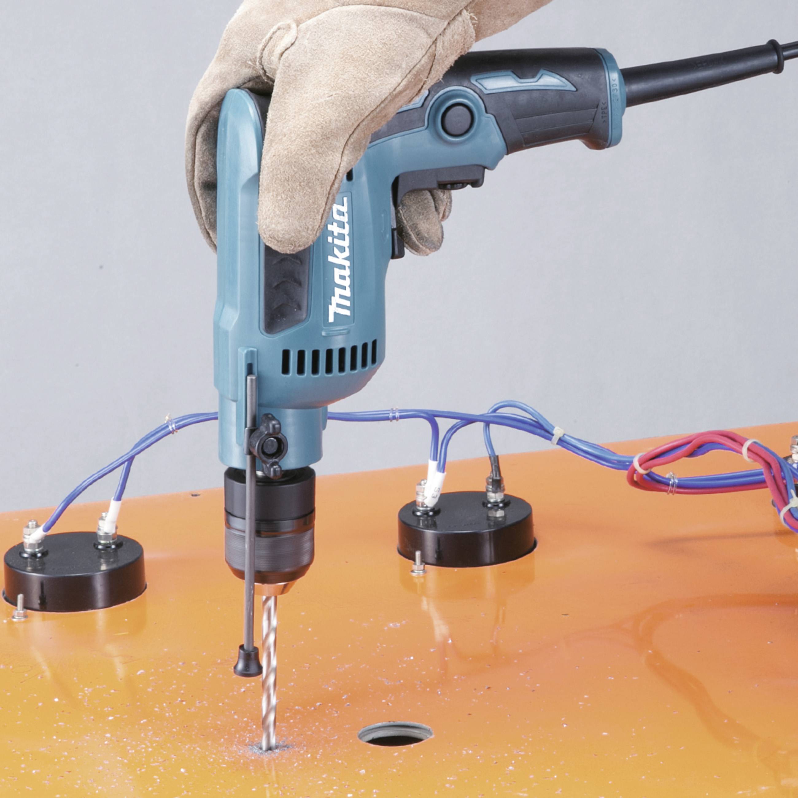 Eine Person bohrt mit einem blauen, elektrischen Bohrer in eine orangefarbene Oberfläche. Schutzhandschuhe sind zu sehen.