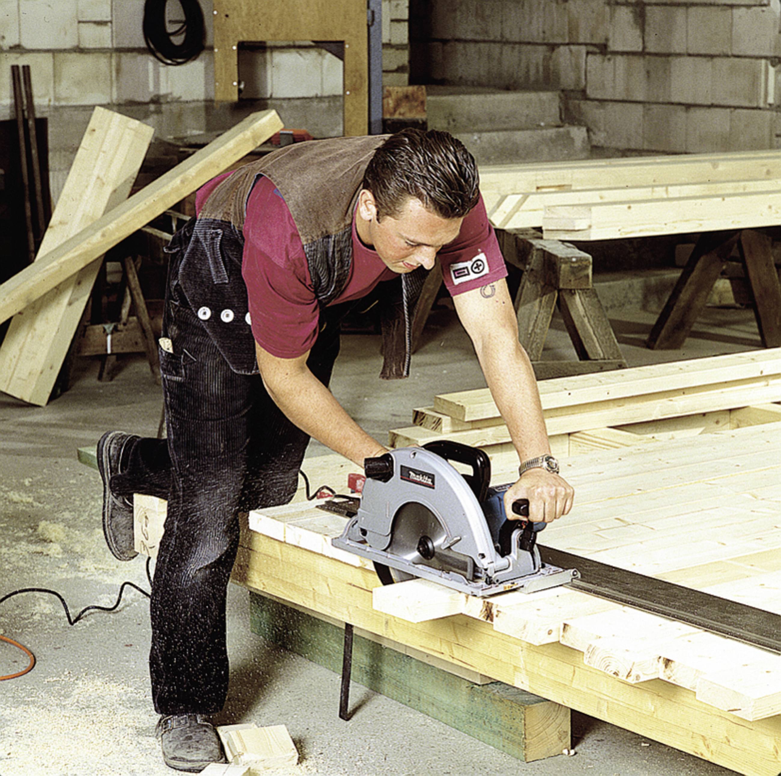 Ein Mann schneidet mit einer elektrischen Kreissäge Holz in einer Werkstatt. Arbeitskleidung und Holzbretter sind im Hintergrund sichtbar.