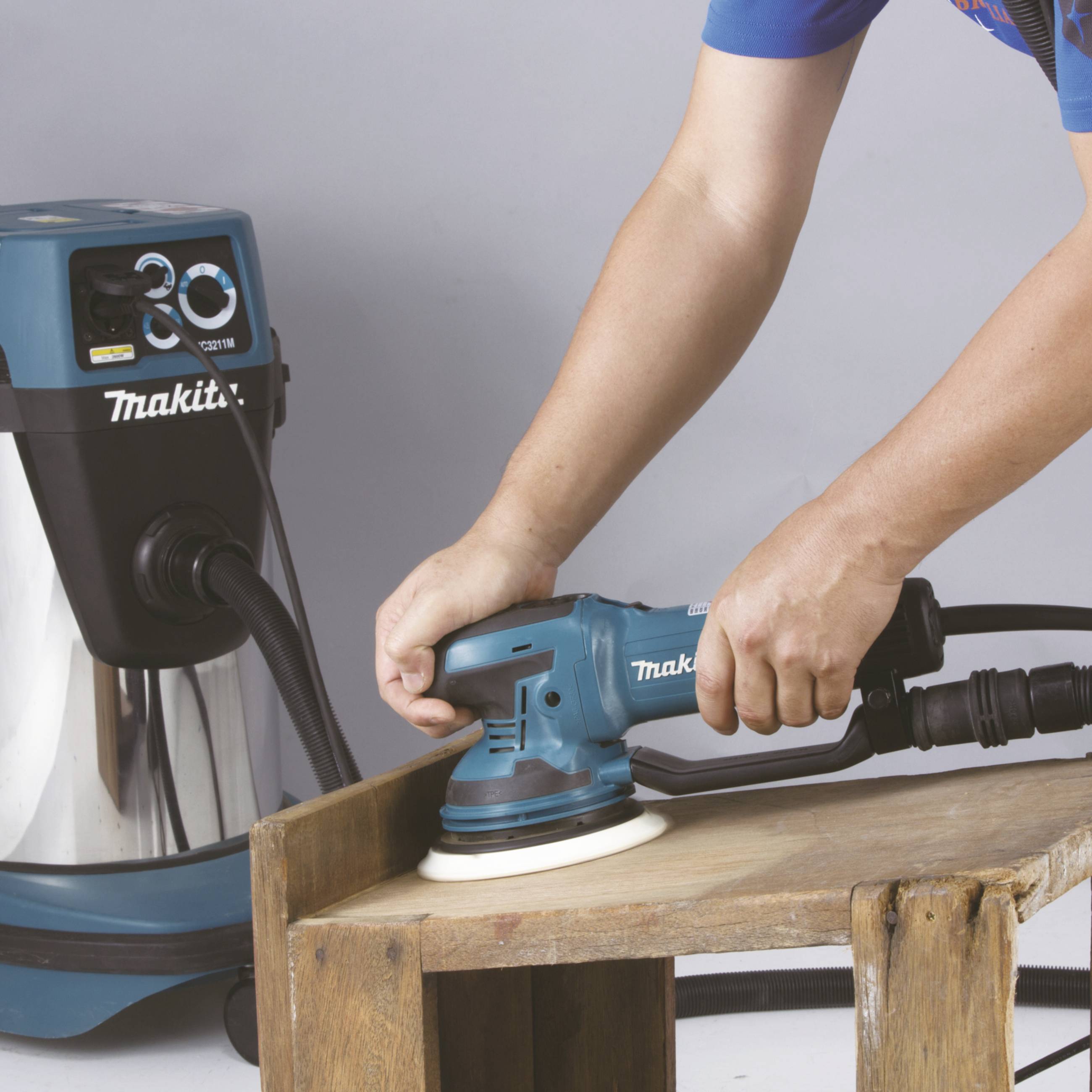 Eine Person schleift mit einem blauen elektrischen Schleifgerät einen Holztisch. Im Hintergrund steht ein blauer Staubsauger.